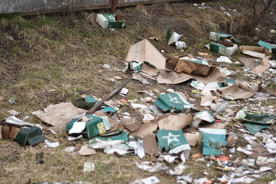 A narrow urban alleyway cluttered with a large black and grey waste collection trolley positioned in the foreground, surrounded by scattered debris and dirt on the ground. In the background, there are